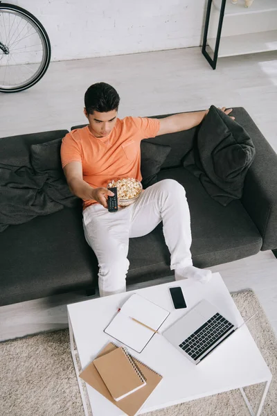 Alto ángulo vista de joven asiático hombre sentado en sofá con bowl de palomitas de maíz y viendo tv — Stock Photo