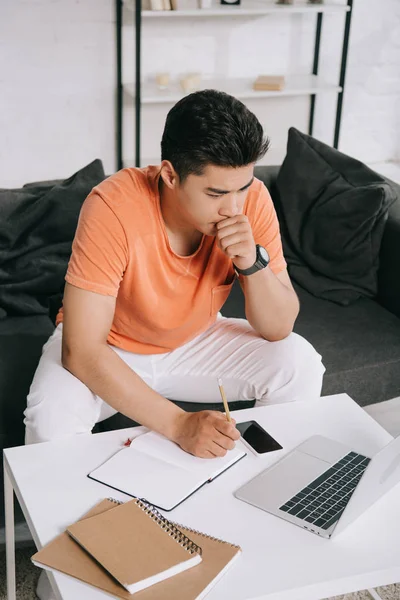 Nachdenklicher asiatischer Mann schaut auf Laptop und schreibt zu Hause in Notizbuch — Stockfoto