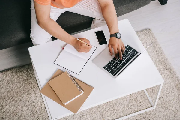 Vista recortada de hombre joven usando el ordenador portátil y la escritura en el cuaderno mientras está sentado en el sofá cerca del escritorio - foto de stock