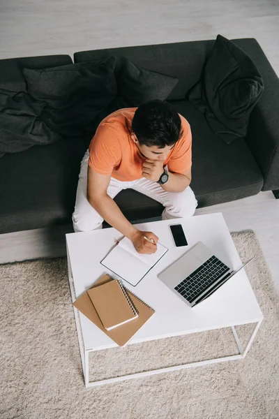 Vue aérienne du jeune homme écrivant dans un cahier et utilisant un ordinateur portable assis sur un canapé près du bureau — Photo de stock