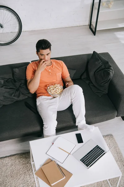 Vista ad alto angolo di giovane uomo asiatico mangiare popcorn mentre seduto sul divano vicino scrivania con computer portatile e notebook — Foto stock