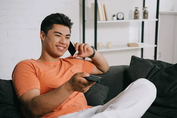 Handsome asian man talking on smartphone and watching tv while sitting on sofa at home — Stock Photo