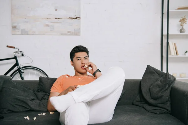Young asian man eating popcorn and looking at camera while sitting on sofa at home — Stock Photo