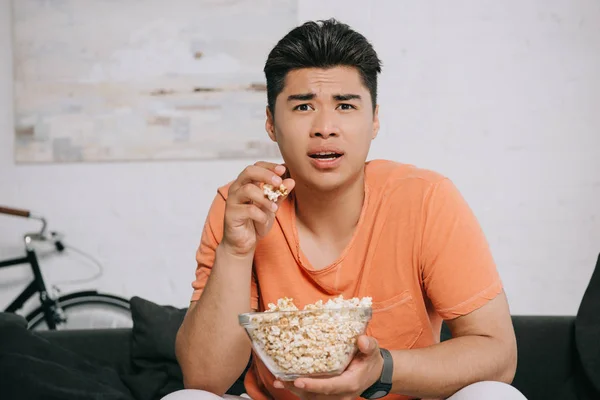 Schockierter asiatischer Mann isst Popcorn und schaut fern, während er zu Hause auf dem Sofa sitzt — Stockfoto