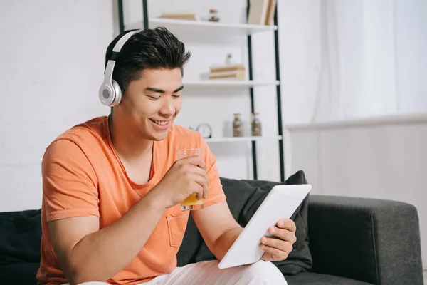 Alegre asiático hombre beber naranja jugo mientras usando digital tablet y escuchar música en auriculares - foto de stock