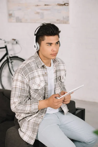 Стомлений азіатський чоловік дивиться геть, слухаючи музику в навушниках і тримаючи цифровий планшет — стокове фото