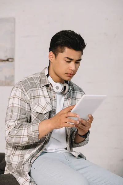 Homme asiatique réfléchi utilisant tablette numérique tout en étant assis dans le canapé à la maison — Photo de stock