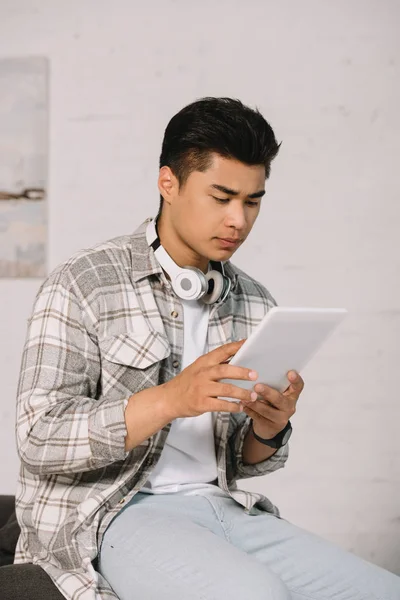 Pensativo asiático homem usando digital tablet enquanto sentado no sofá em casa — Fotografia de Stock