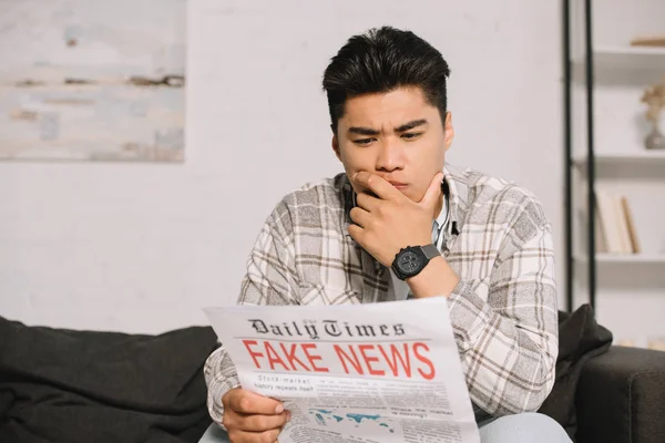 Серйозний азіатський чоловік читає фальшиву газету, сидячи на дивані вдома — стокове фото