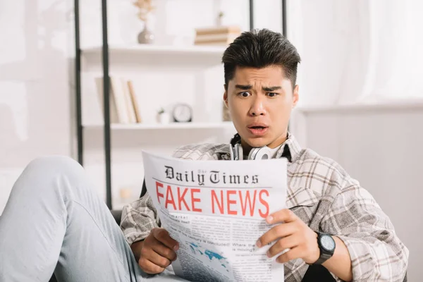 Шокований азіатський чоловік читає фальшиву газету, сидячи на дивані вдома — стокове фото