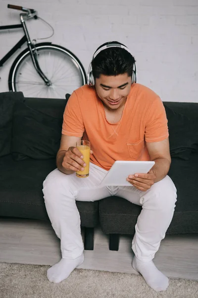 Sonriente asiático hombre bebiendo jugo de naranja y usando digital tablet mientras sentado en sofá y escuchar música en auriculares - foto de stock