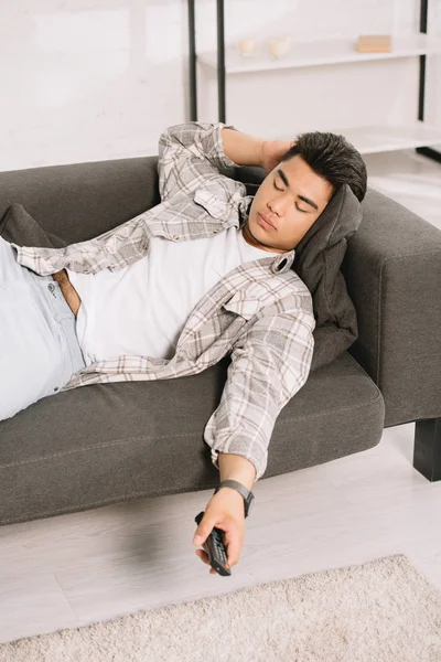 Exhausted asian man sleeping on couch while holding tv remote controller — Stock Photo