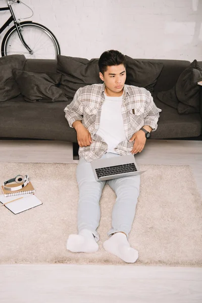 Overhead view of thoughtful asian man sitting on floor with laptop and looking away — Stock Photo