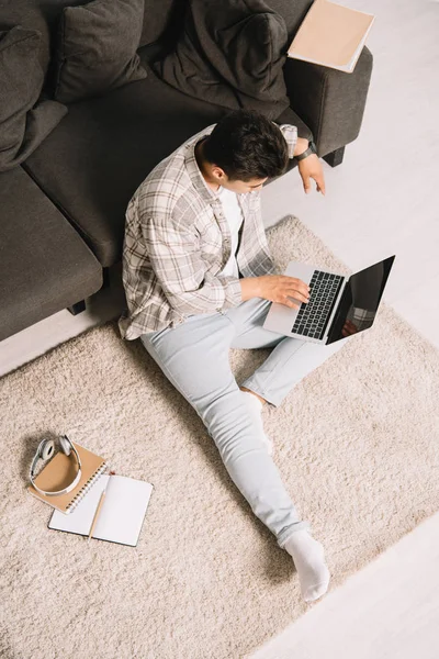 Вид сверху на молодого человека, сидящего дома на полу и использующего ноутбук — стоковое фото
