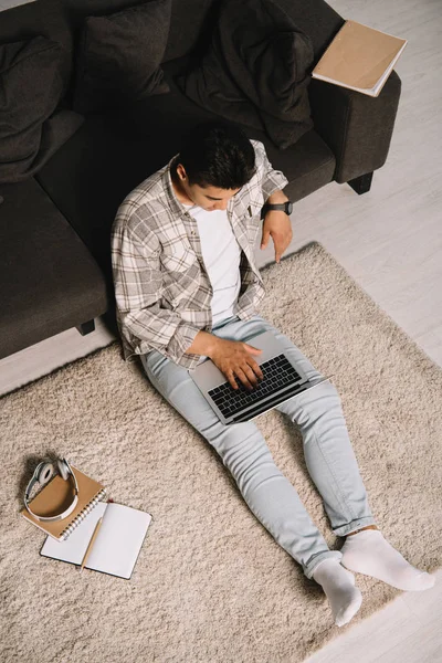 Vue aérienne du jeune homme assis sur le sol près du canapé et utilisant un ordinateur portable à la maison — Photo de stock