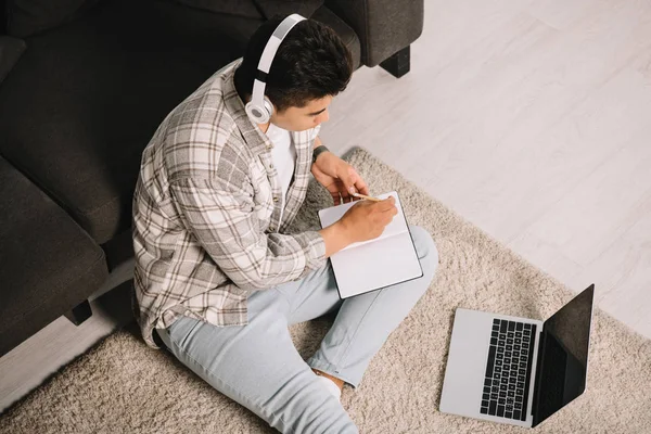 Visão aérea do jovem em fones de ouvido sentado no chão perto do laptop e escrevendo no notebook — Fotografia de Stock