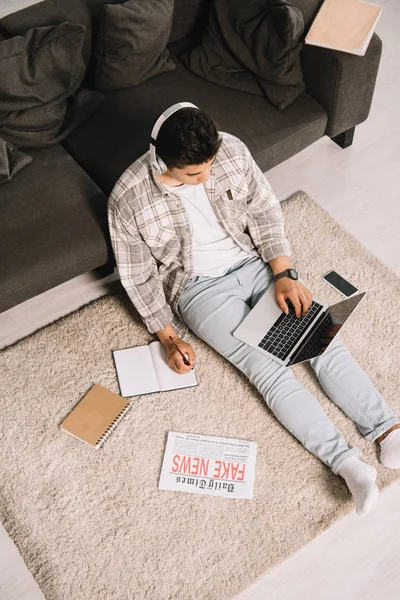 Overhead view of young man in headphones sitting on floor at home, using laptop and writing in notebook — Stock Photo