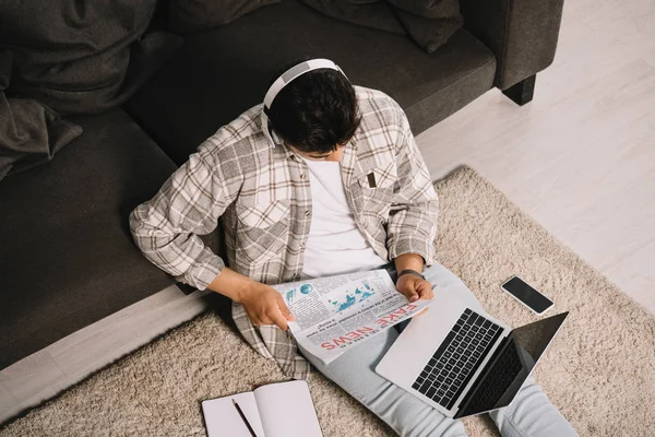 Vue aérienne du jeune homme lisant de fausses nouvelles journal tout en étant assis sur le sol avec ordinateur portable — Photo de stock