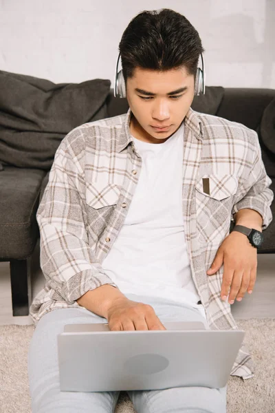 Attentif asiatique homme dans casque à l'aide d'ordinateur portable tout en étant assis sur le sol à la maison — Photo de stock