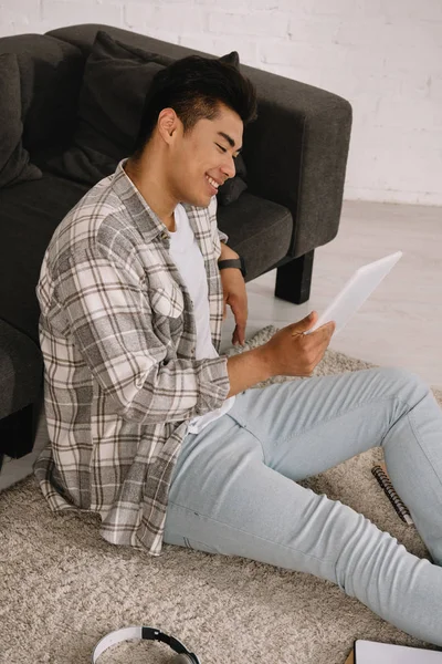 Alegre asiático homem usando digital tablet enquanto sentado no chão perto de sofá em casa — Fotografia de Stock