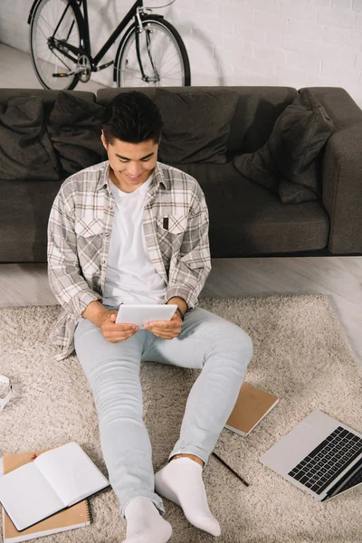 Вид зверху усміхнений азіатський чоловік сидить на підлозі біля дивана і використовує цифровий планшет — стокове фото