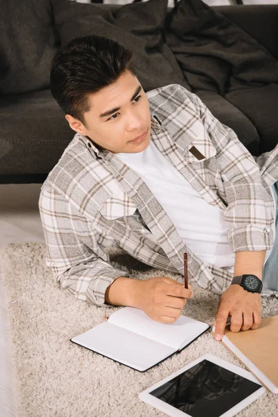 Pensive asian man looking away while lying on floor near notebook and digital tablet with blank screen — Stock Photo