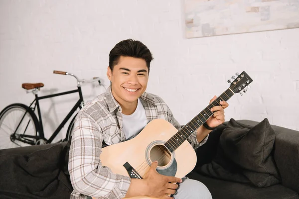 Fröhlicher asiatischer Mann spielt Akustikgitarre und lächelt in die Kamera — Stockfoto