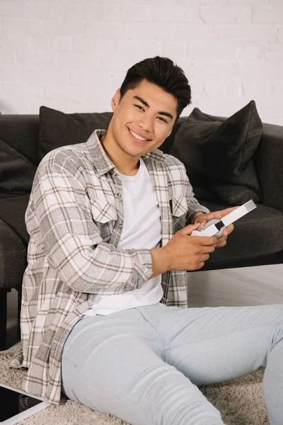 Cheerful asian man smiling at camera while sitting on floor at home and holding headphones — Stock Photo