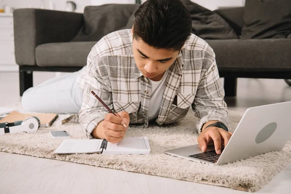 Aufmerksamer asiatischer Mann schreibt in Notizbuch und benutzt Laptop, während er zu Hause auf dem Boden liegt — Stockfoto