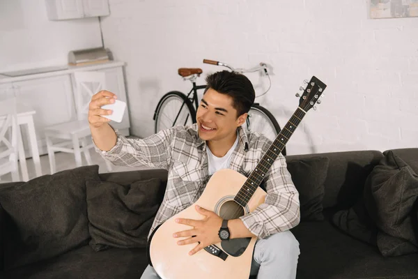 Веселий азіатський чоловік бере селфі зі смартфоном, сидячи на дивані з акустичною гітарою — стокове фото