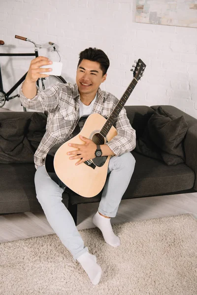 Усміхнений азіатський чоловік бере селфі зі смартфоном, сидячи на дивані з акустичною гітарою — стокове фото