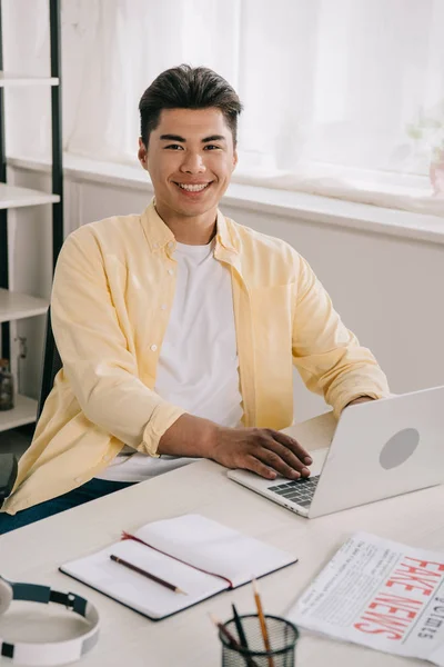Allegro asiatico uomo sorridente a fotocamera mentre seduta a scrivania e utilizzando laptop — Foto stock