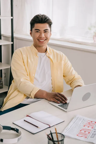 Joyeux asiatique homme souriant à la caméra tout en étant assis au bureau et en utilisant ordinateur portable — Photo de stock