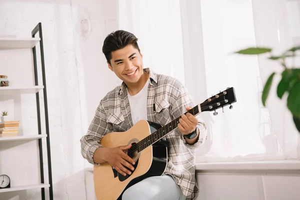 Handsome, young asian man looking away while playing acoustic guitar at home — Stock Photo