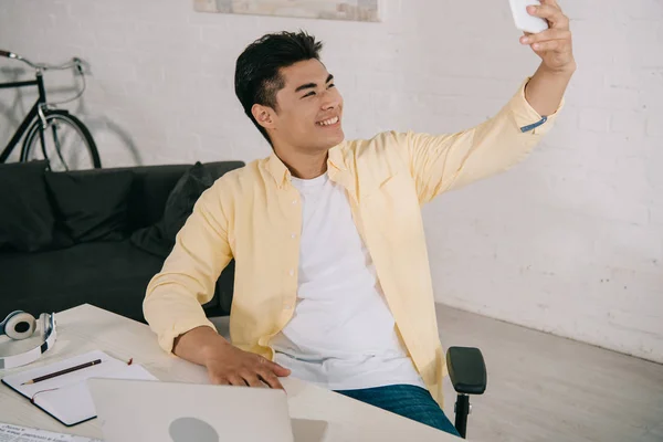 Sonriente asiático hombre tomando selfie con smartphone mientras sentado en escritorio en casa - foto de stock