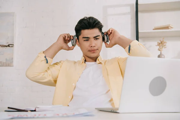 Thoughtful asian man in headphones sitting at desk and looking at laptop — Stock Photo
