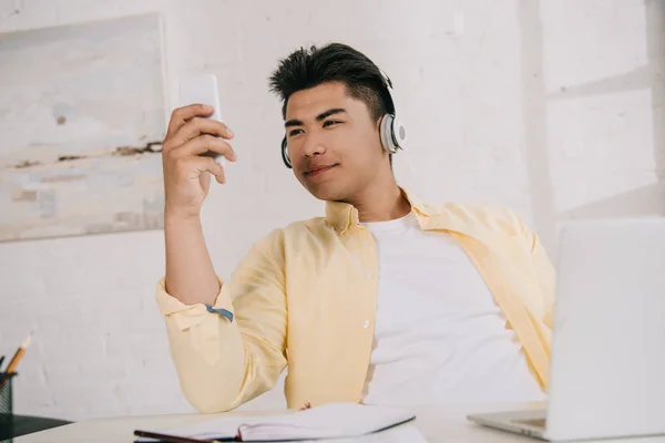 Joven, sonriente asiático hombre en auriculares tomando selfie con smartphone - foto de stock