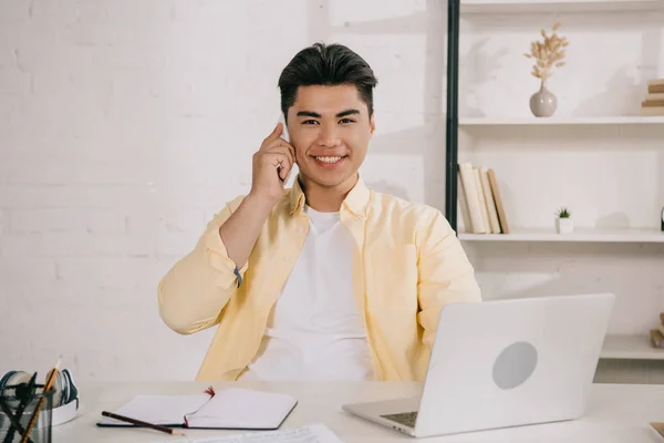 Guapo asiático hombre hablando en smartphone y sonriendo a cámara mientras sentado en el lugar de trabajo en la oficina - foto de stock