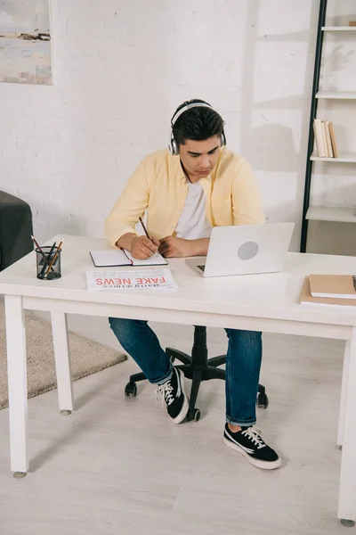 Focalizzato asiatico uomo in cuffie guardando laptop mentre scrive in notebook — Foto stock