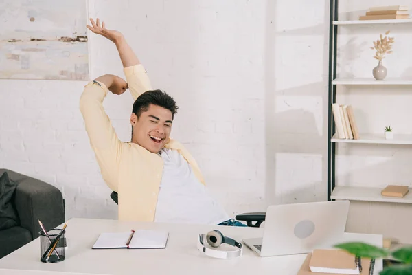 Joven asiático hombre estiramiento y sonriendo mientras sentado en escritorio cerca de portátil - foto de stock