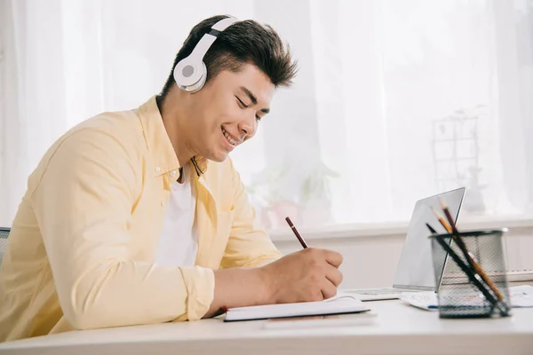 Alegre asiático hombre en auriculares escritura en notebook mientras sentado en escritorio - foto de stock