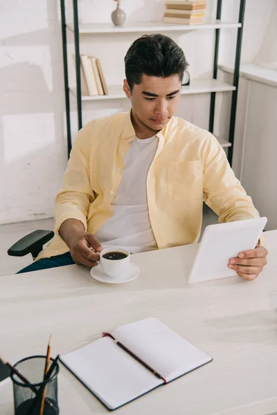 Handsome asian man holding cup of coffee while sitting at desk and using digital tablet — Stock Photo