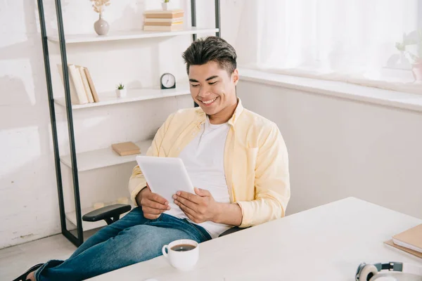 Cheerful asian man looking at digital tablet while sitting at desk near cup of coffee — Stock Photo