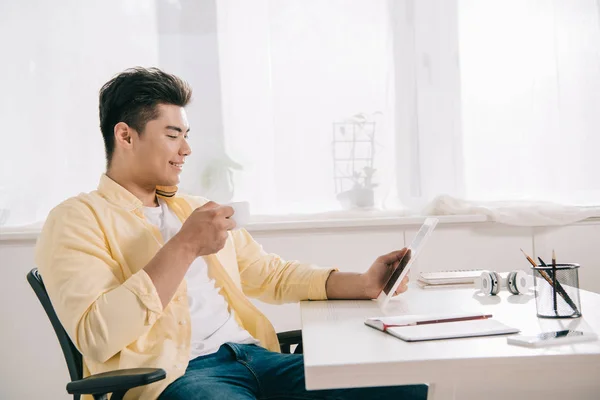 Junger asiatischer Mann benutzt digitales Tablet und trinkt Kaffee, während er am Tisch sitzt — Stockfoto