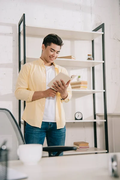 Selettivo fuoco di sorridente asiatico uomo lettura libro mentre in piedi vicino rack a casa — Foto stock