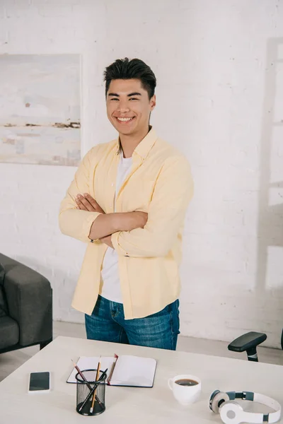 Junger, fröhlicher asiatischer Mann, der zu Hause am Schreibtisch steht und in die Kamera lächelt — Stockfoto