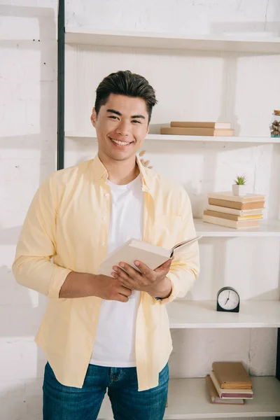 Joven, alegre asiático hombre sosteniendo libro mientras de pie cerca rack y sonriendo a la cámara - foto de stock