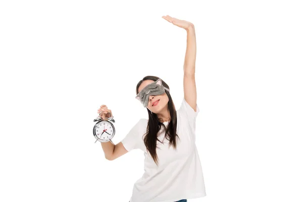 Brunette young woman in cat sleeping eye mask outstretching herself and holding alarm clock isolated on white — Stock Photo