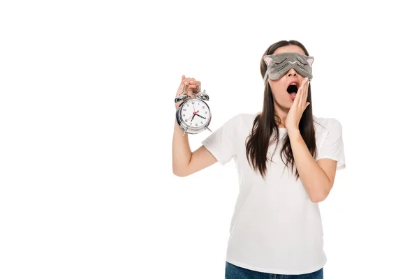 Brunette young woman in cat sleeping eye mask yawning and holding alarm clock isolated on white — Stock Photo