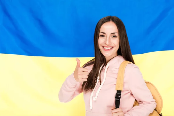 Усміхнена брюнетка молода жінка з рюкзаком показує великий палець на фоні українського прапора — стокове фото